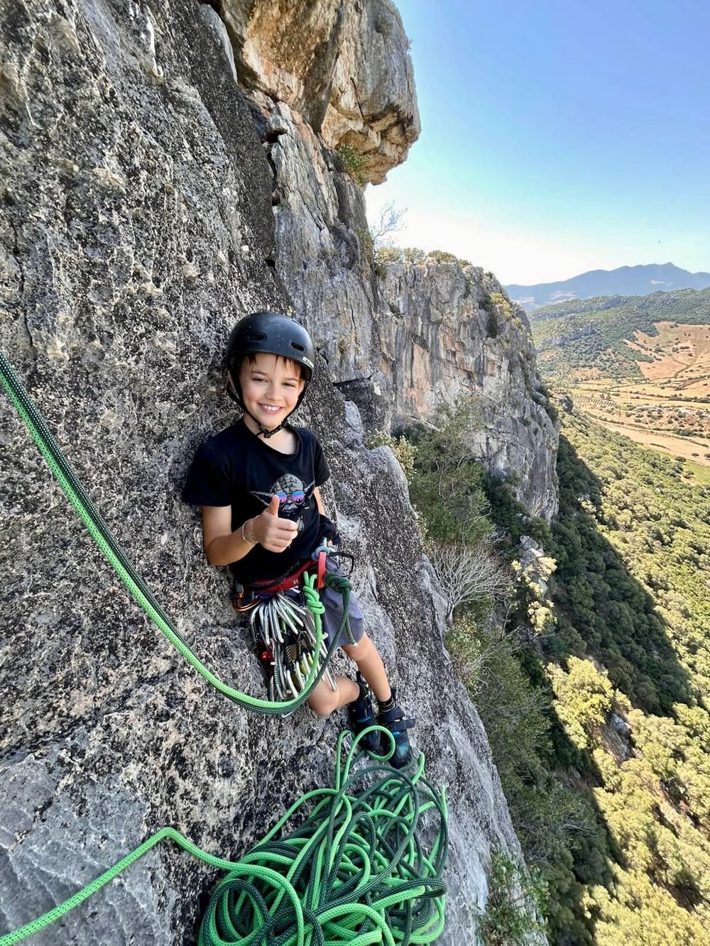 Liam klettert lächelnd mit Helm, grünem Seil und sichtbarer Kletterausrüstung am Kalksteinfels auf Sardinien, mit Daumen hoch vor grünen Hügeln, Feldern und Bergen
