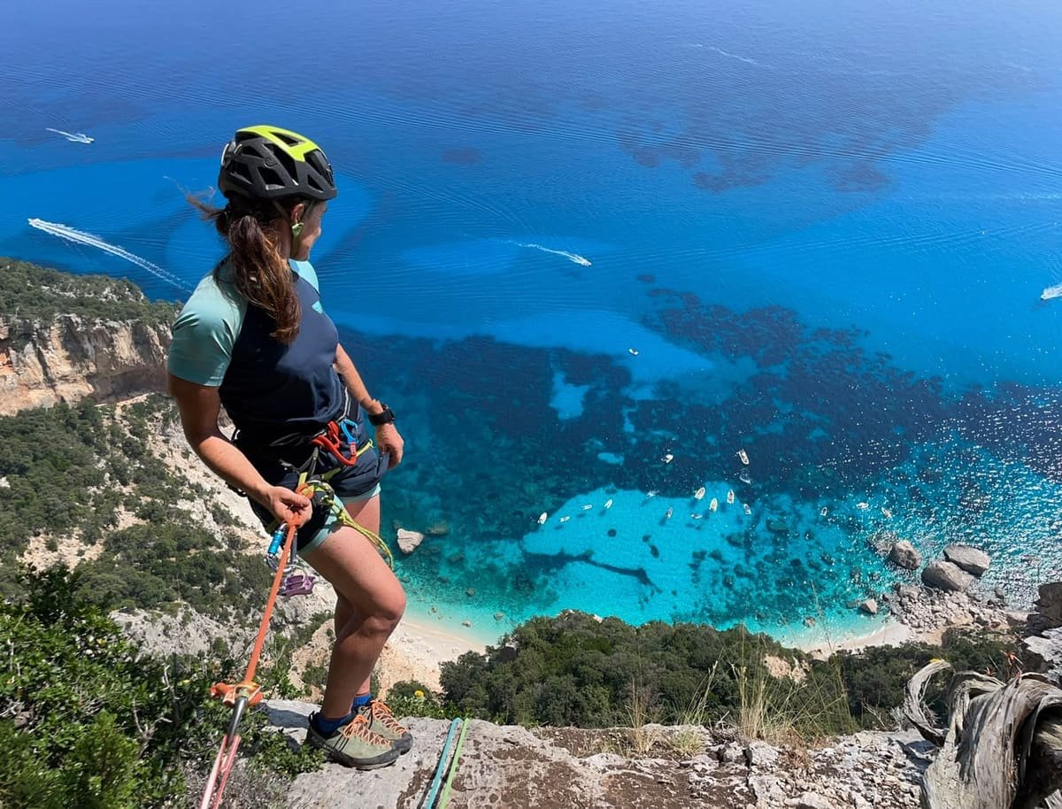 Kletterin am Abseilstand über einer türkisblauen Bucht, Blick von oben auf Boote und Strand.
